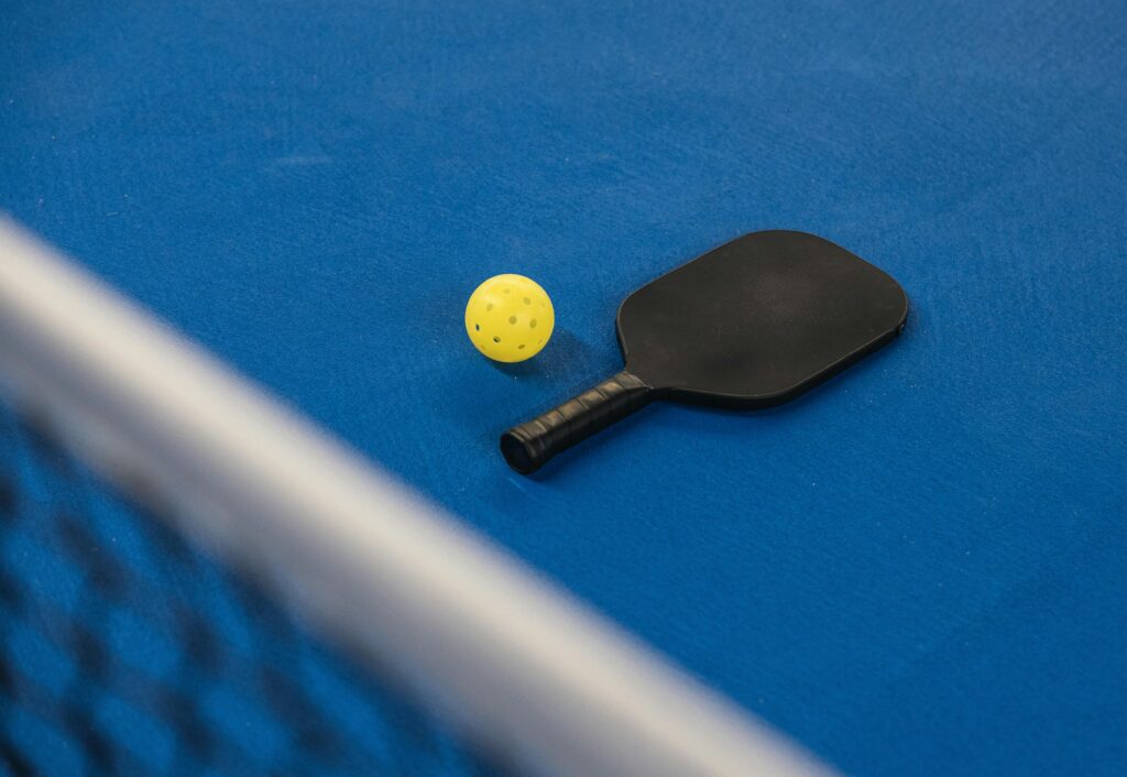 A pickleball paddle and ball resting on a blue court surface with a net in focus.